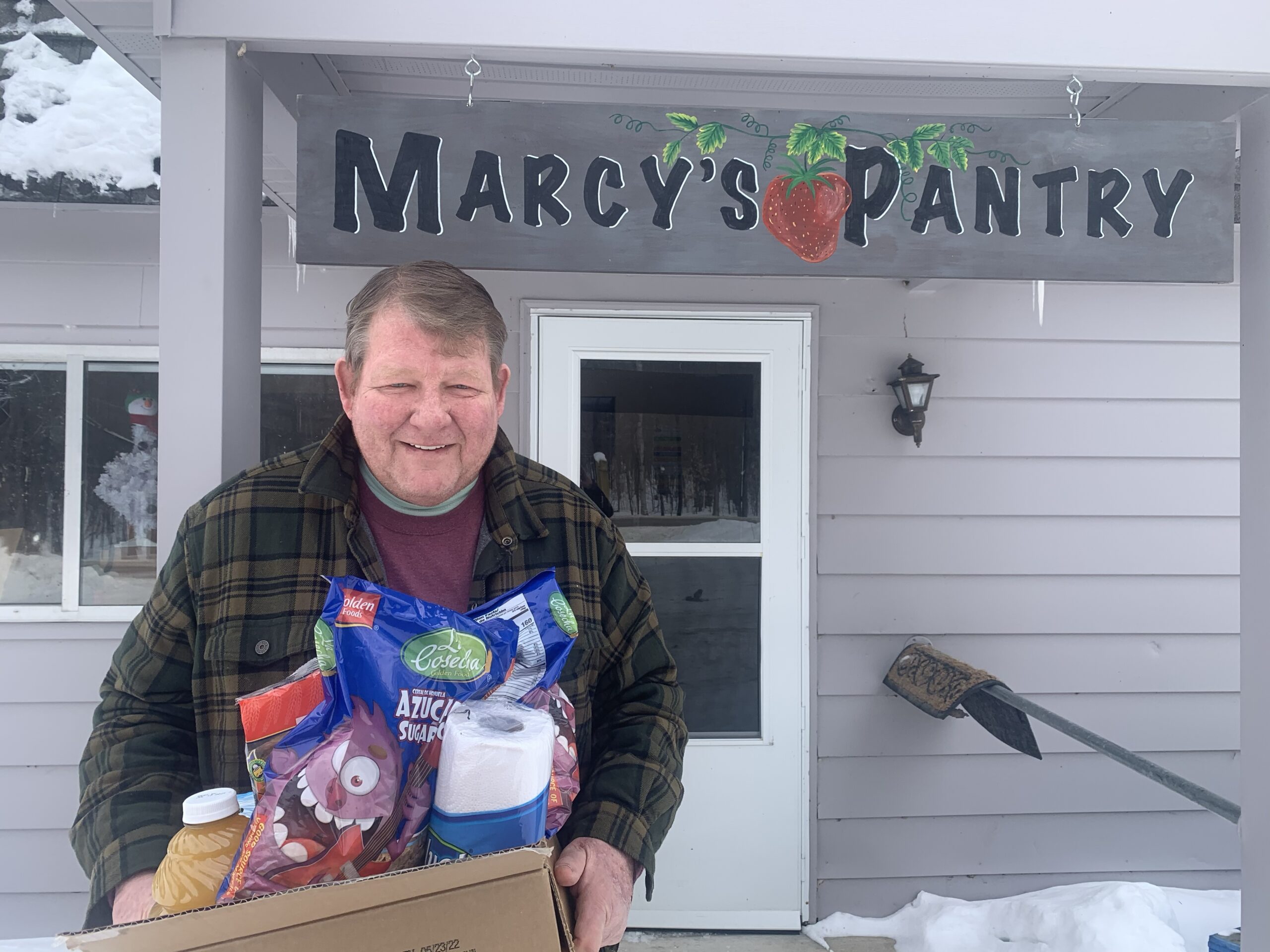 Stu holds a box of food and other items in front of a Marcy's Pantry handpainted sign. The sign includes a strawberry.